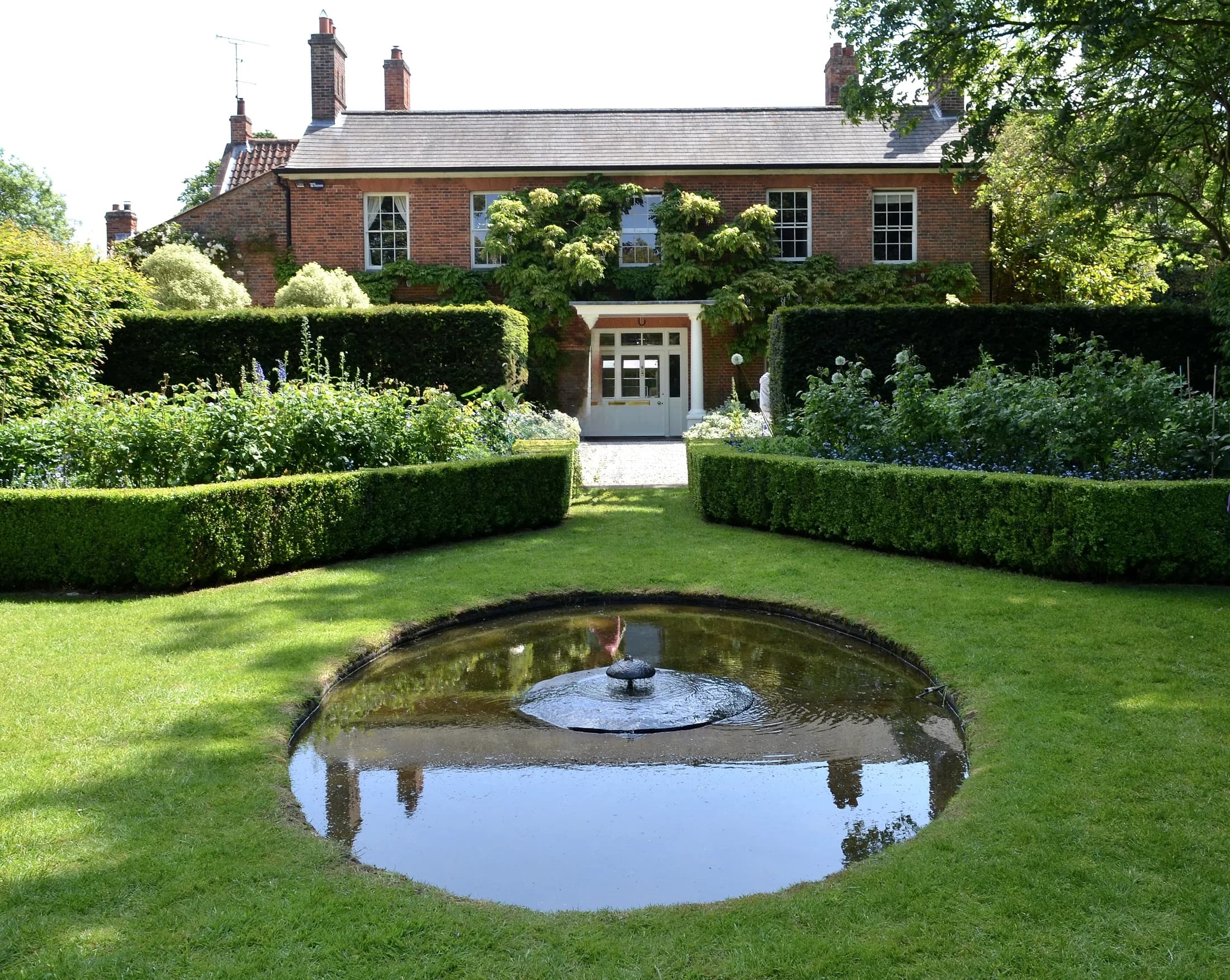 A red-brick period house behind clipped hedges and a still garden pond.