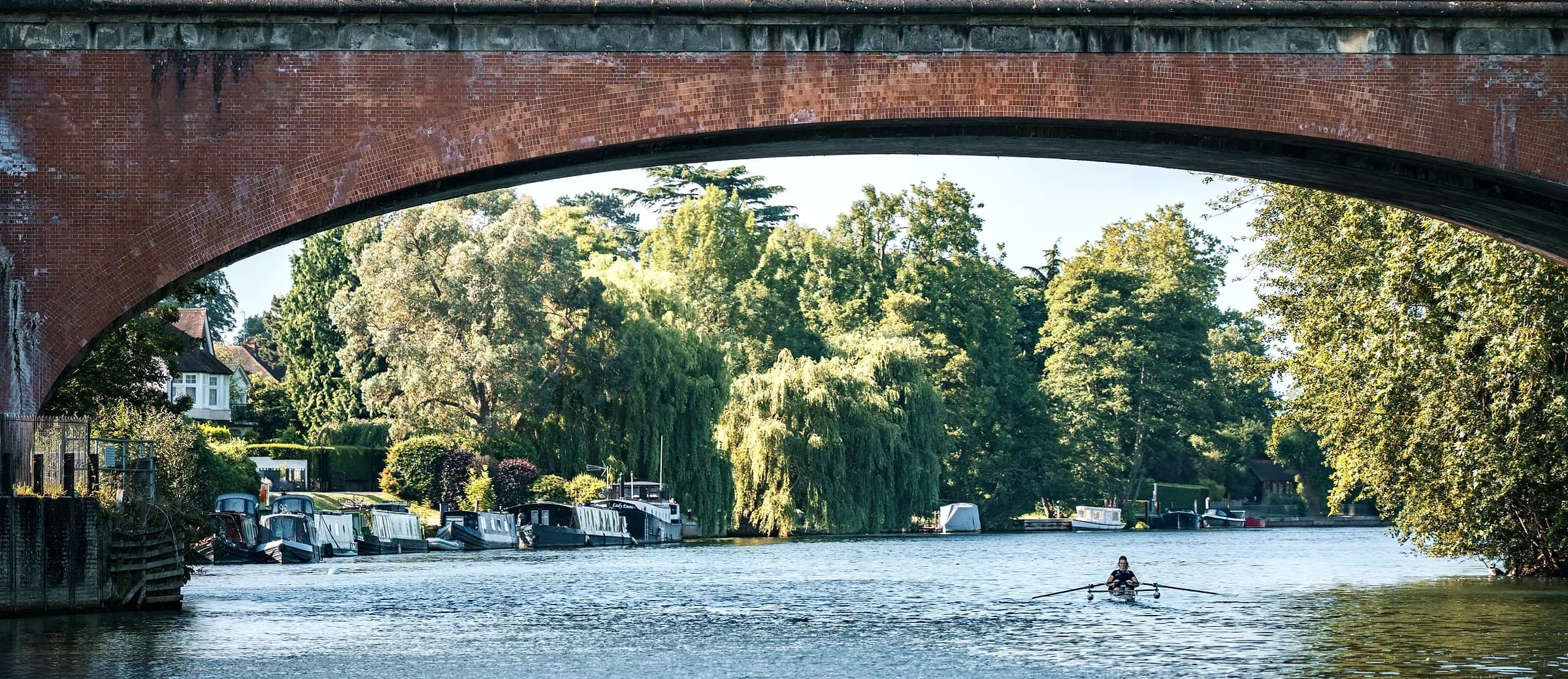 A sculler on the Thames below a brick railway arch, with moored boats and willows along the bank.